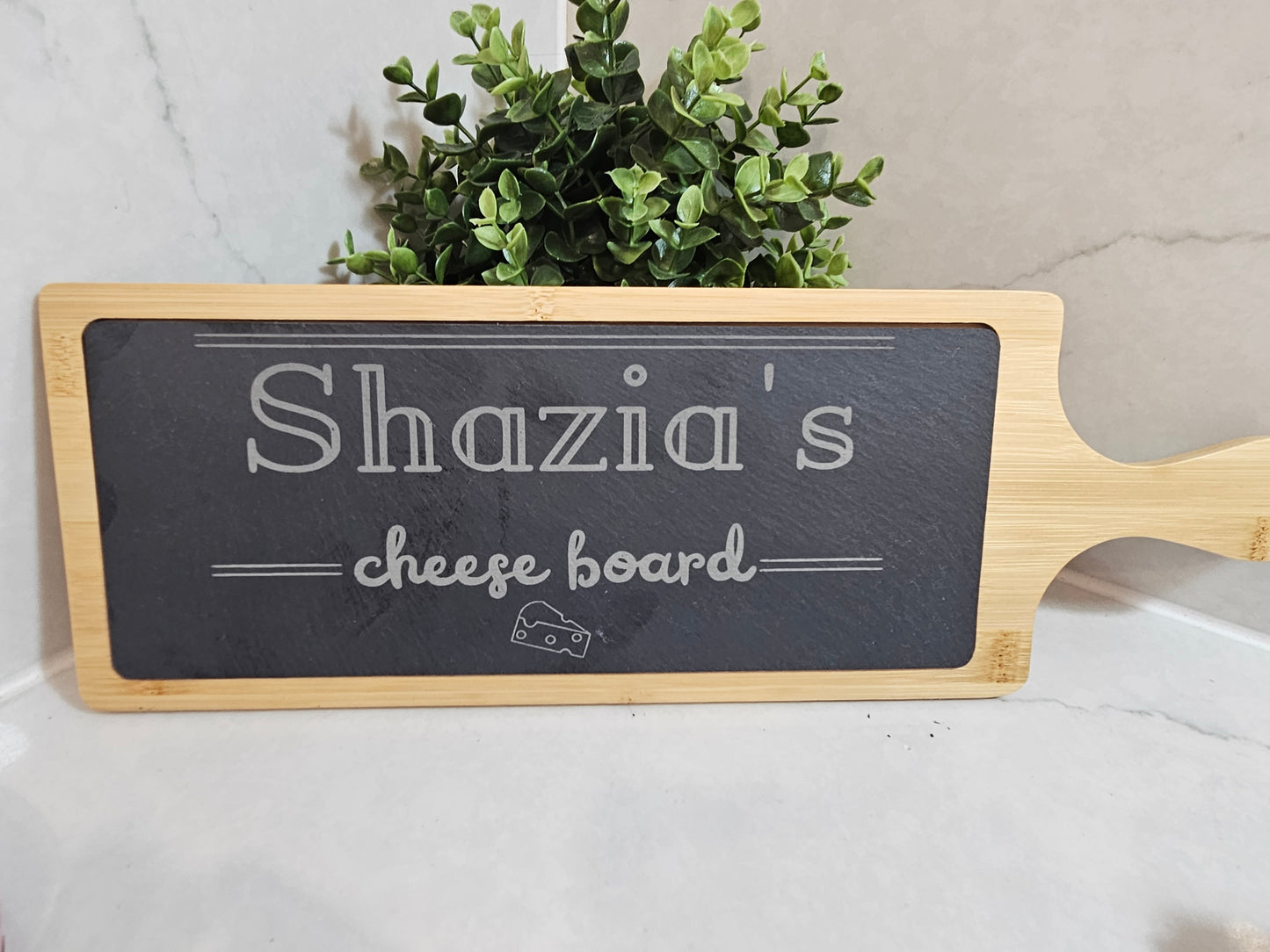 Personalised cheese board with 'Shazia's cheese board' text on a marble surface with a plant in the background.