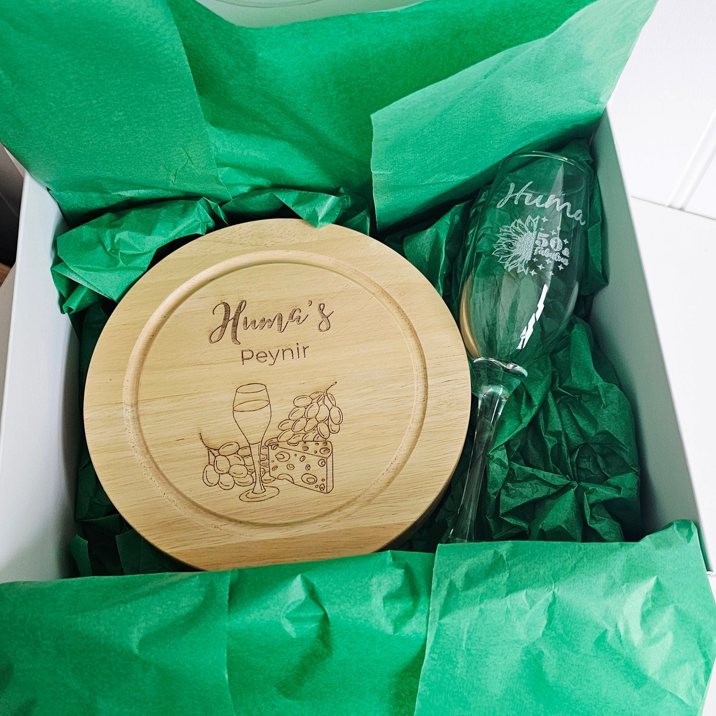 Wooden cheese board and glass with 'Huma's Peynir' branding on green tissue paper.