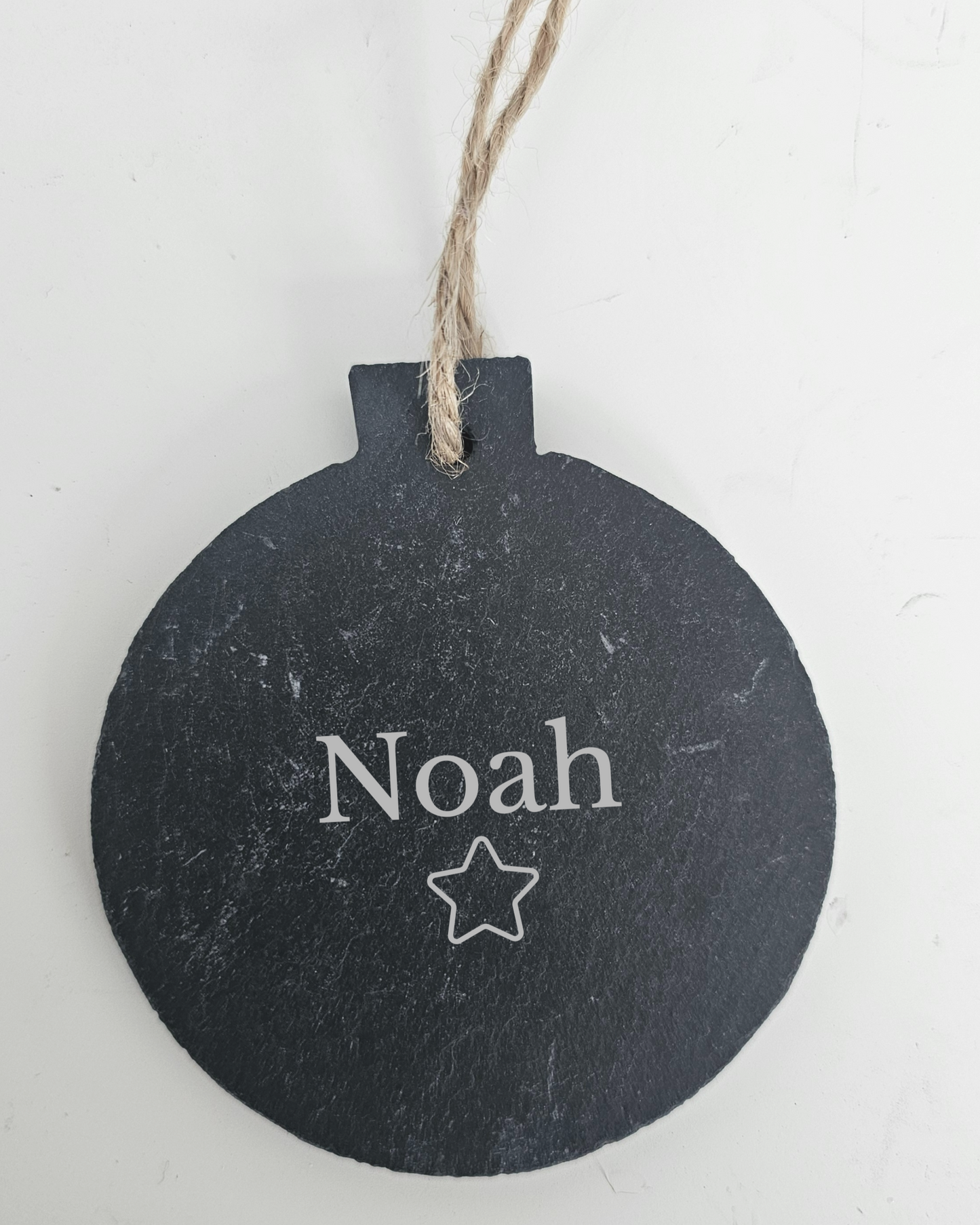 Black round slate bauble personalised with 'Noah' and a star engraved on it, hanging on a white background.
