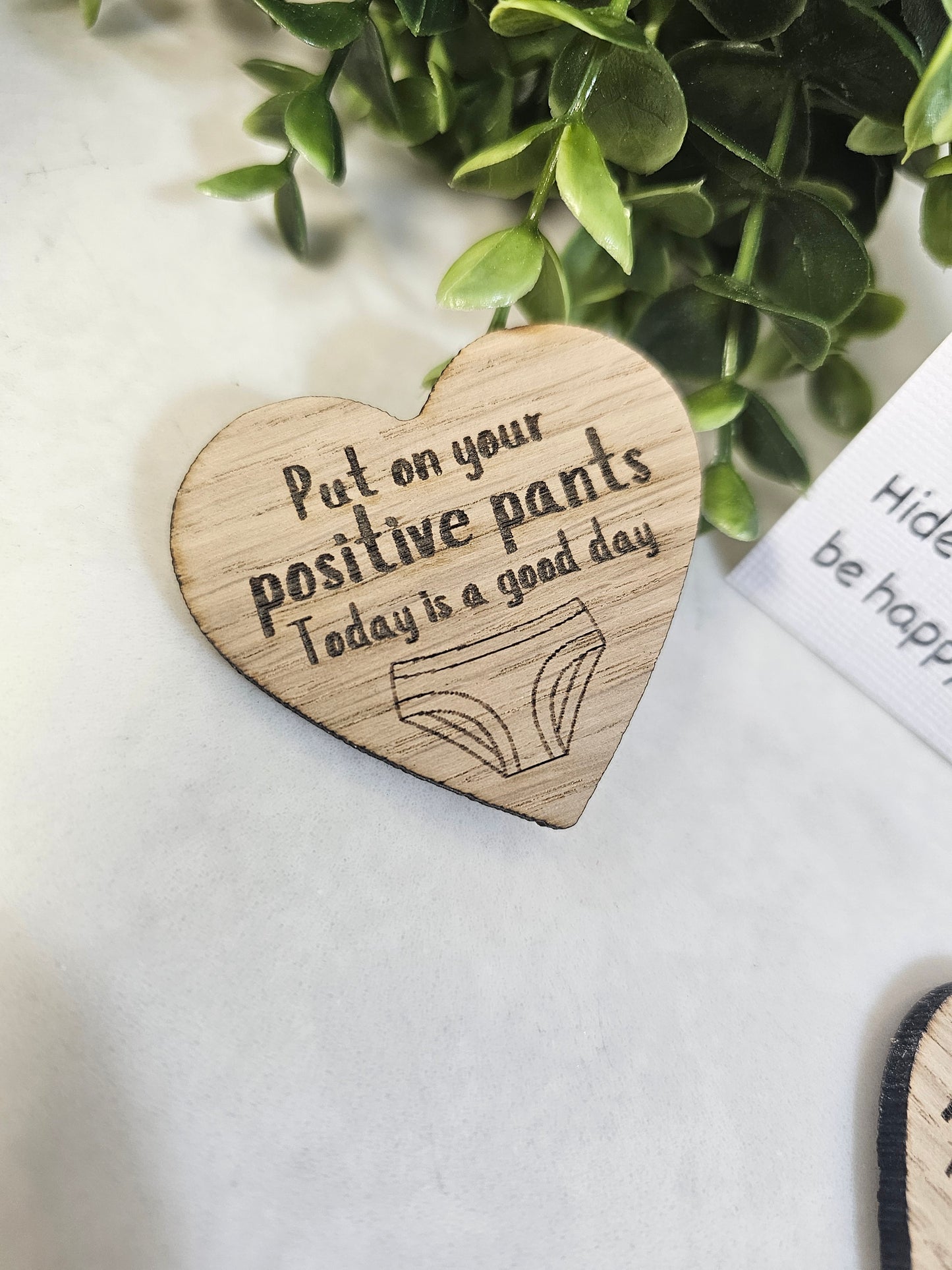 Wooden heart-shaped plaque with motivational quote on a white surface with greenery.