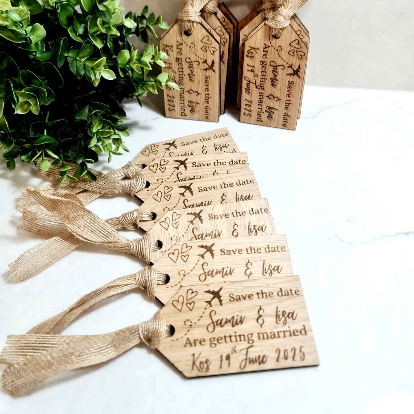 A set of wooden luggage tag-shaped save the date magnets with engraved text, tied with a string to a greenery decoration in the background.