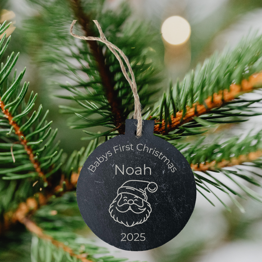 Black ornament with 'Baby's First Christmas' text on a Christmas tree branch.