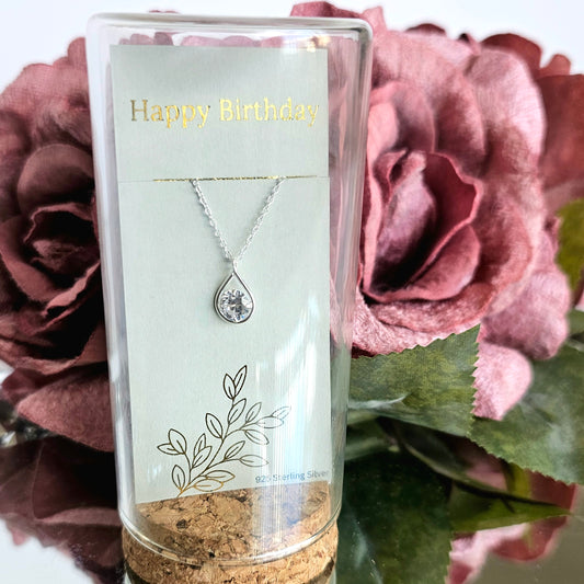 A sterling silver pear-shaped necklace with cubic zirconia, presented in a glass jar with a cork stopper, placed in front of a floral background.