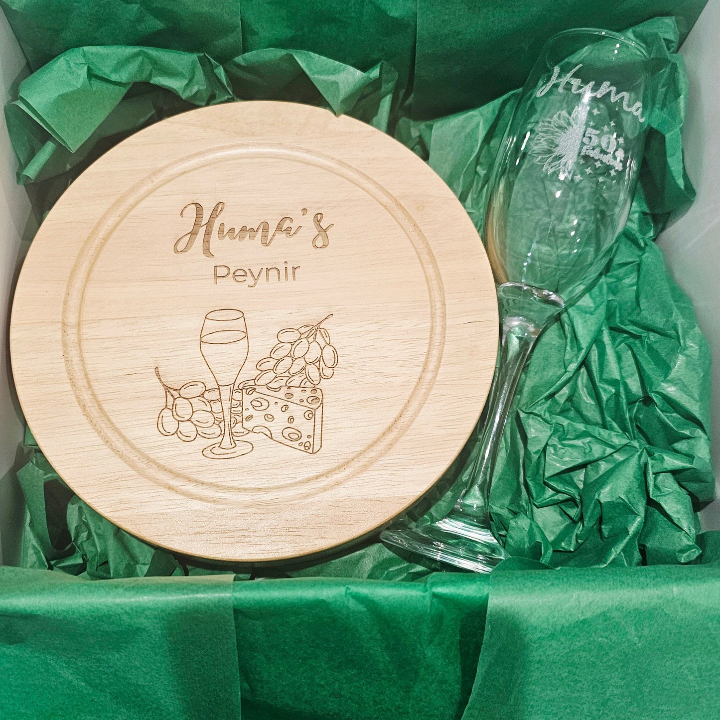 Wooden cheese board with engraved design and text, accompanied by two glasses on green tissue paper.