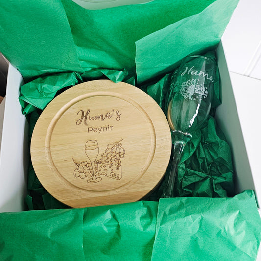 Wooden cheese board and glass with 'Huma's Peynir' branding on green tissue paper.