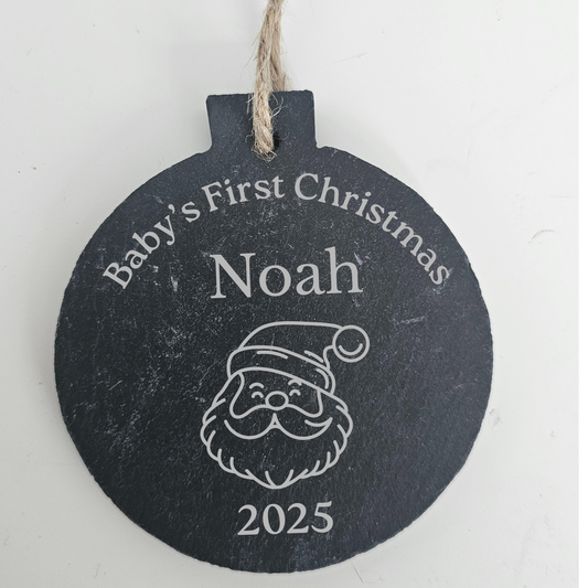 Round slate ornament with Santa Claus design and text 'Baby's First Christmas Noah 2025' on a white background.