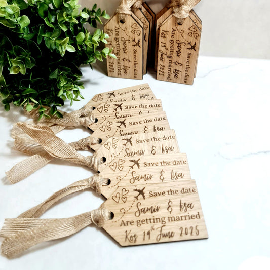 A set of wooden luggage tag-shaped save the date magnets with engraved text, tied with a string to a greenery decoration in the background.